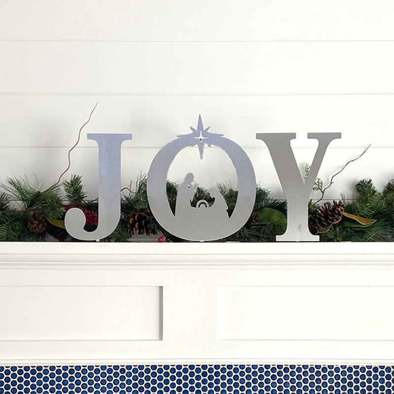Metal letters J O Y with manger scene silhouette inside the o, standing on mantel in front of green garland.