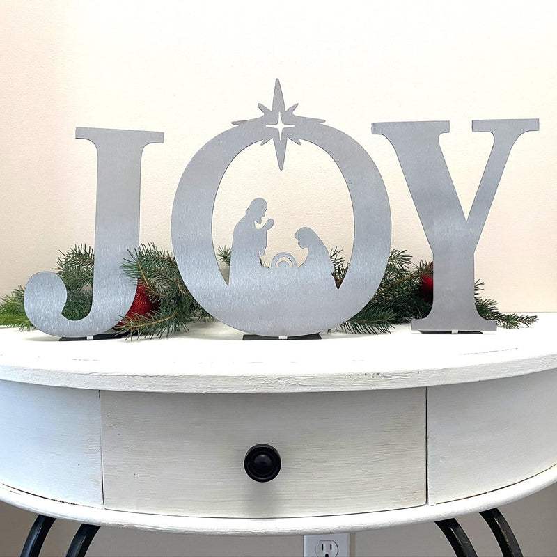 Metal letters J O Y with manger scene silhouette inside the o, standing on shelf in front of greenery.