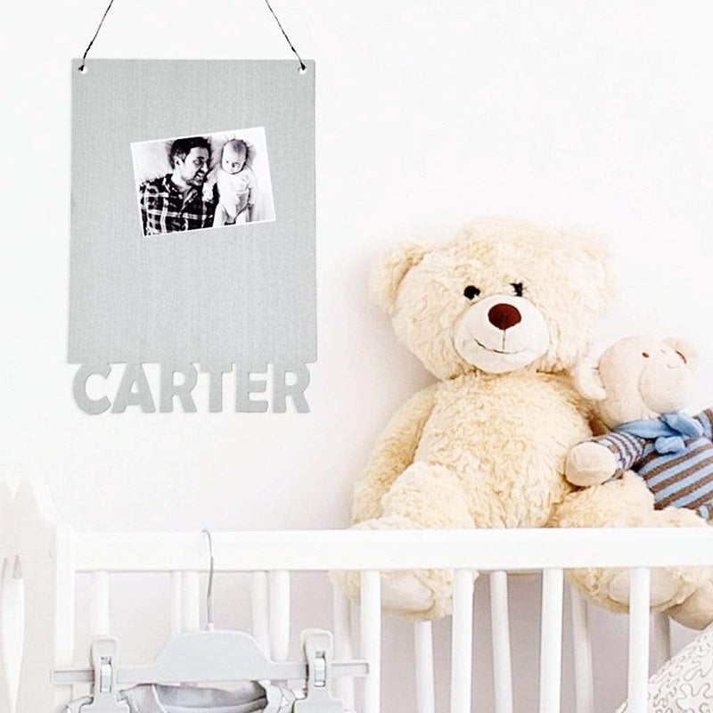 Rectangle magnetic board with name etched at the bottom and photo held on by magnet, hanging on wall above baby crib.