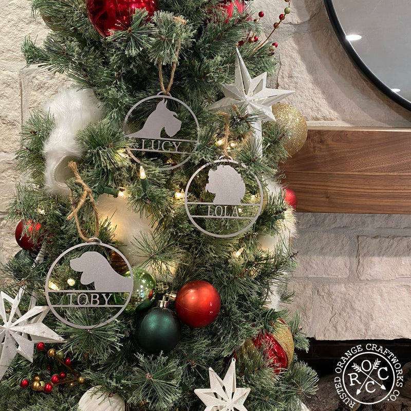 Round metal ornaments with personalized dog name and breed silhouette, hanging on Christmas tree.