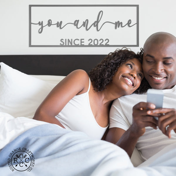 Picture of couple lounging in bed with metal sign above bed that says you and me since 2022