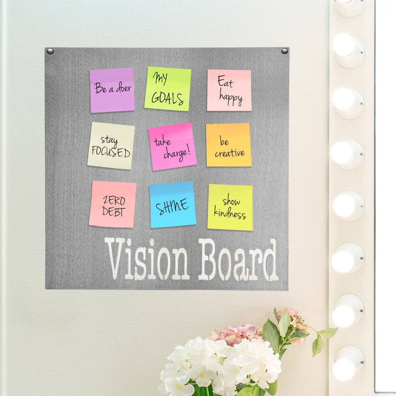 Square metal magnet board with vision board etched at the bottom and post-it notes stuck on it, hanging on the wall above flowers.