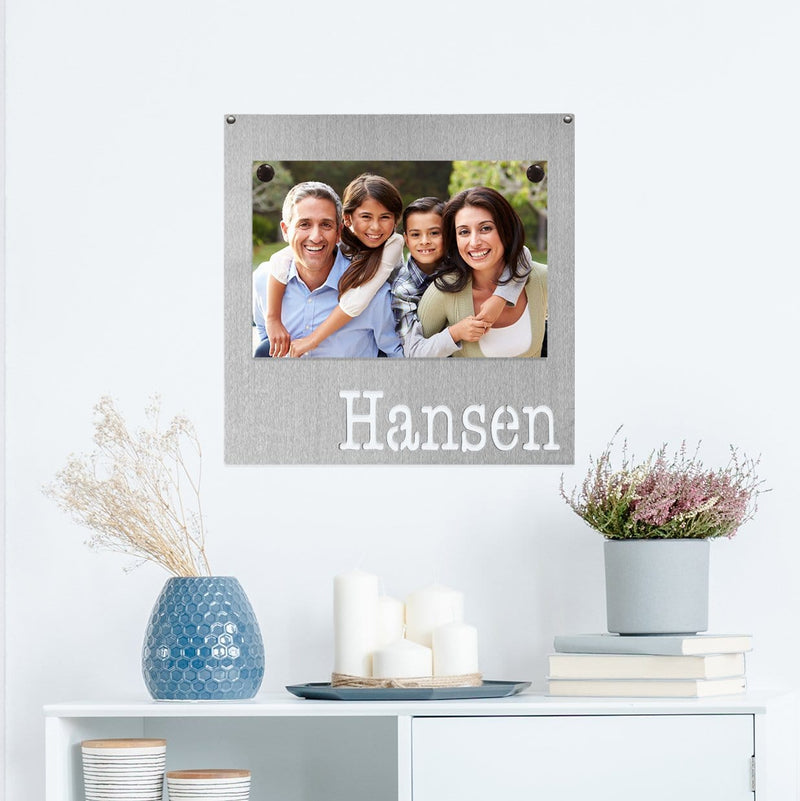 Square metal magnet board with the name Hansen etched at the bottom and family photo stuck on it, hanging on the wall above table.