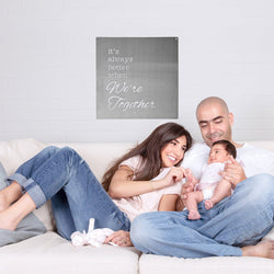 Square metal sign saying it's always better when we're together hanging on wall above smiling family sitting on couch.