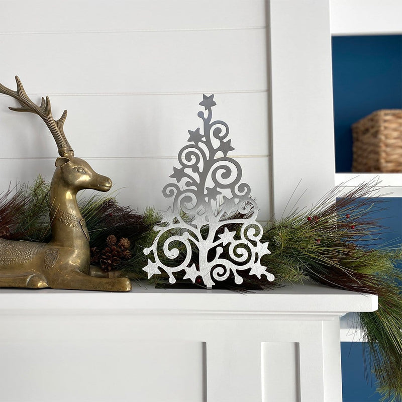 Metal Christmas tree cutout with swirls and stars throughout, standing on mantel with garland and gold reindeer statue. 