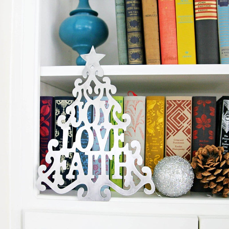 Metal Christmas tree cutout with words joy love faith in the center, standing on shelf with books.