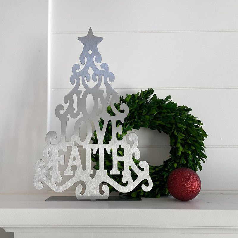 Metal Christmas tree cutout with words joy love faith in the center, standing on shelf next to wreath and Christmas bauble.
