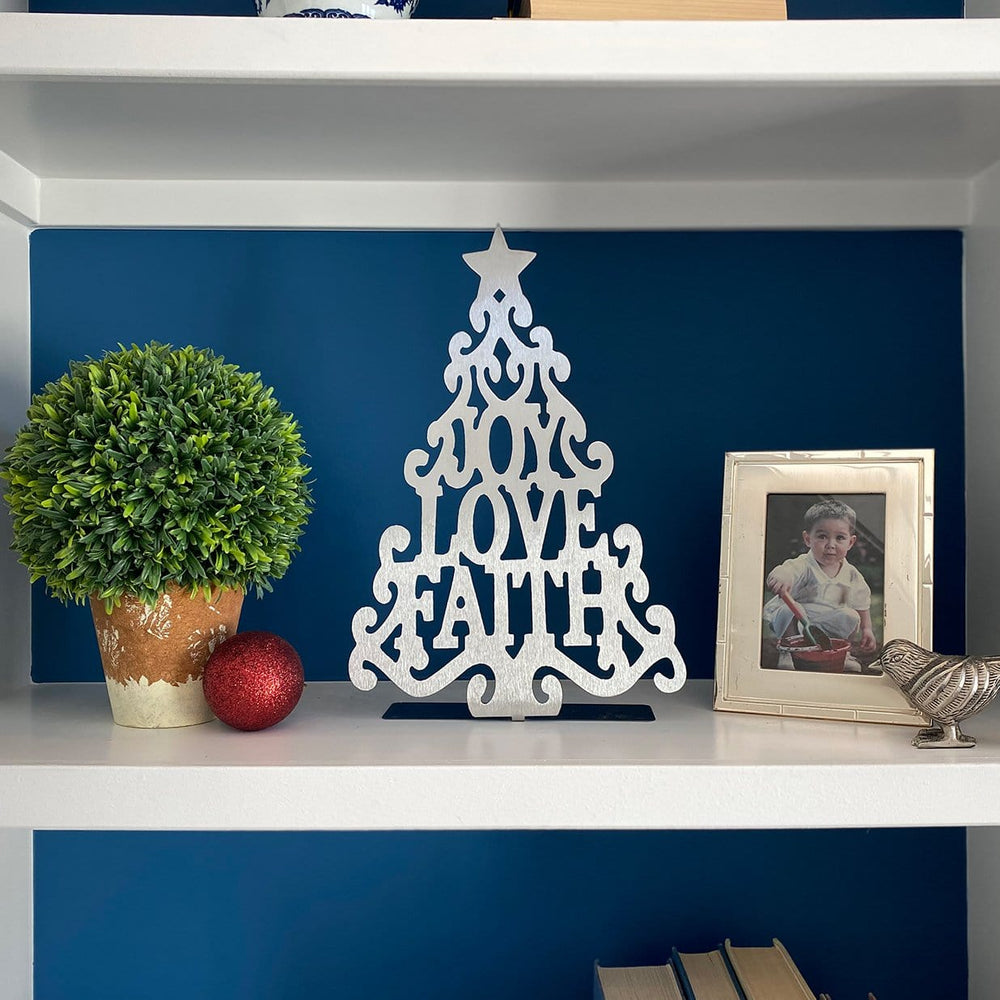 Metal Christmas tree cutout with words joy love faith in the center, standing on shelf next to plant and photo.