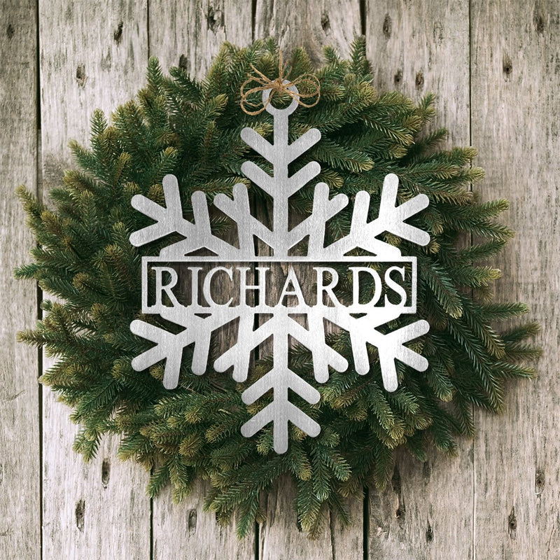 Metal snowflake with name in center hanging with green wreath on wood fence. 