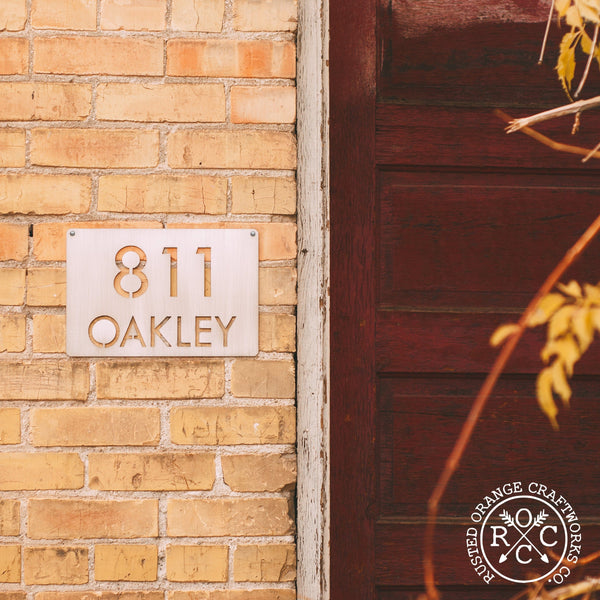Metal rectangle sign with home address hanging outside brick house.