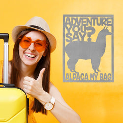 Rectange metal sign with alpaca in center and saying, adventure you say? Alpaca my bag. Hanging on wall next to smiling woman with suitcase.