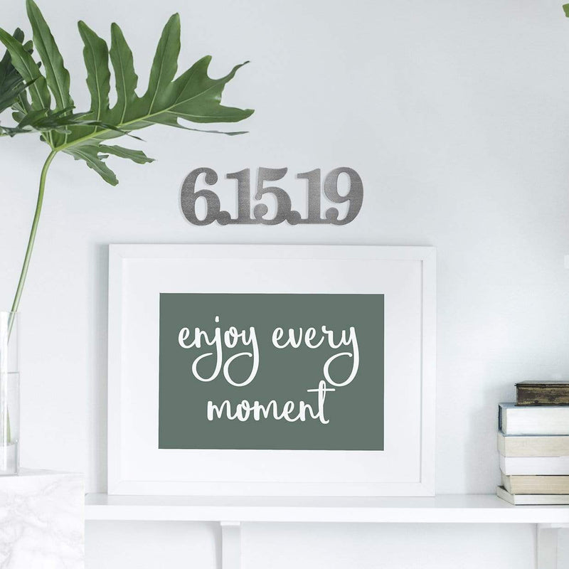 Metal sign with date 6/15/19 hanging on wall above sign saying enjoy every moment.