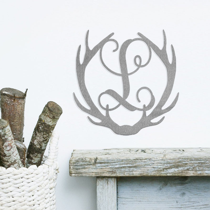 Metal deer antlers with monogram L in the middle, hanging on wall above wooden shelf.
