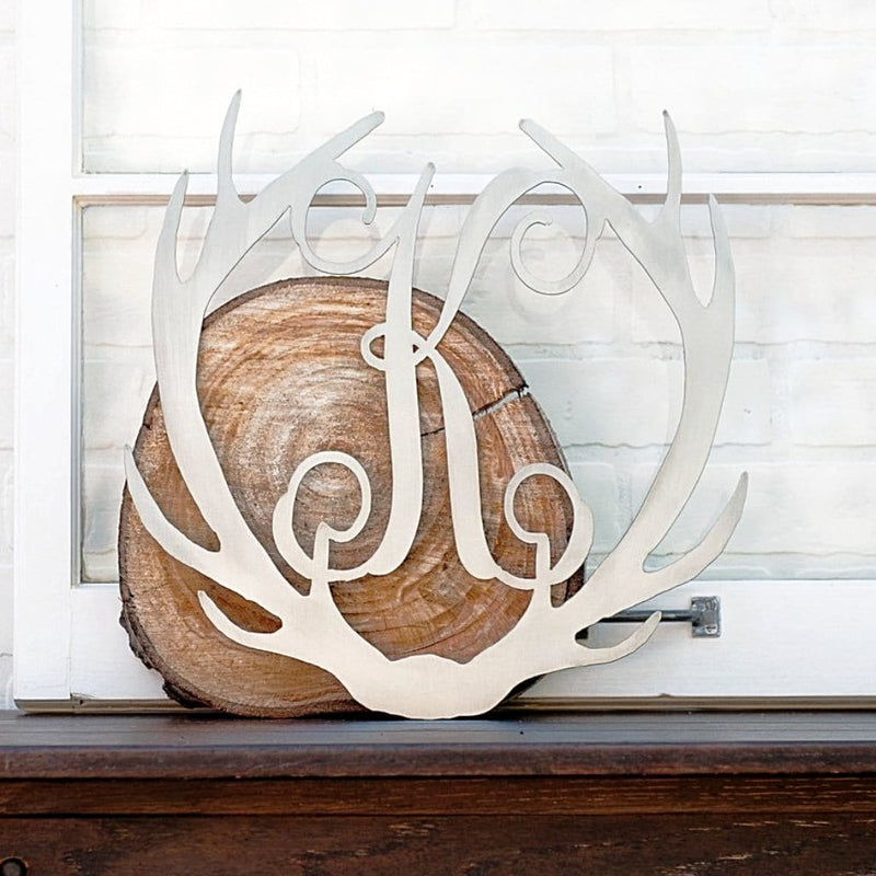 Metal deer antlers with monogram K in the middle, sitting on mantel leaning on piece of wood.