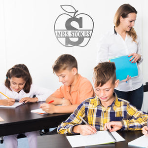 Apple shaped sign with name and monogram in the middle, hanging on classroom wall with smiling teacher and students working at desks.