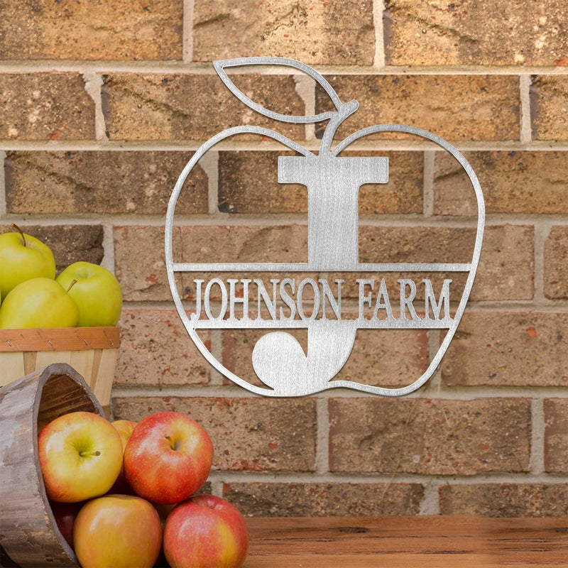 Apple shaped sign with name and monogram in the middle, hanging on brick wall next to buckets of apples.