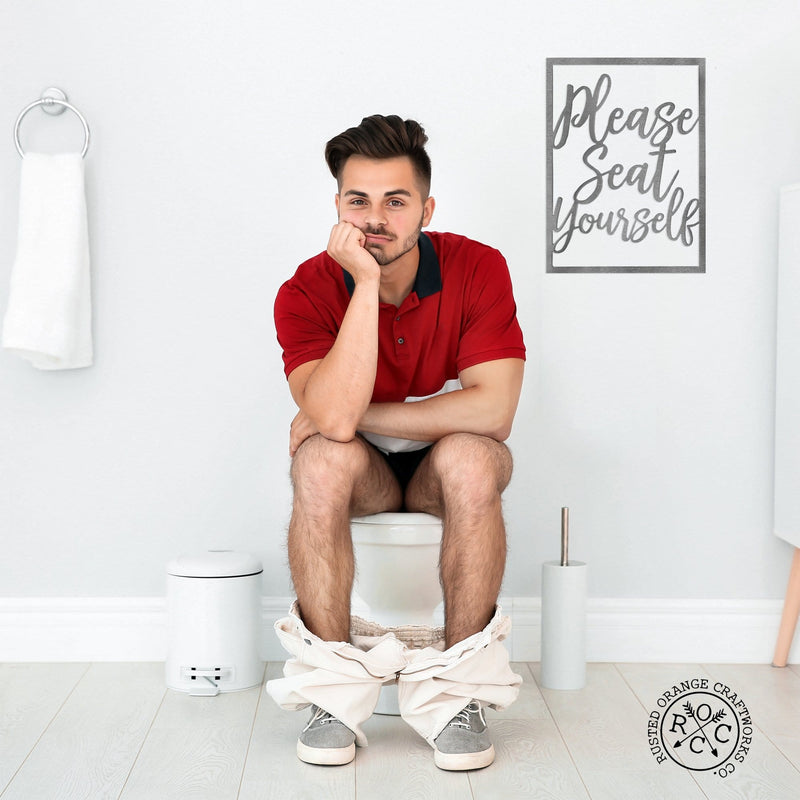 Metal rectangle humorous bathroom phrase sign hanging on wall above man sitting on toilet.