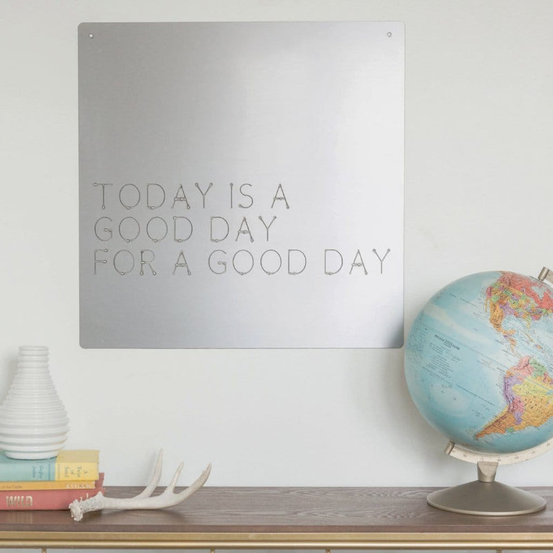 Square metal sign saying today is a good day for a good day, hanging on wall above shelf.
