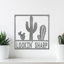 Square metal sign with cactus that says lookin sharp, hanging on wall.