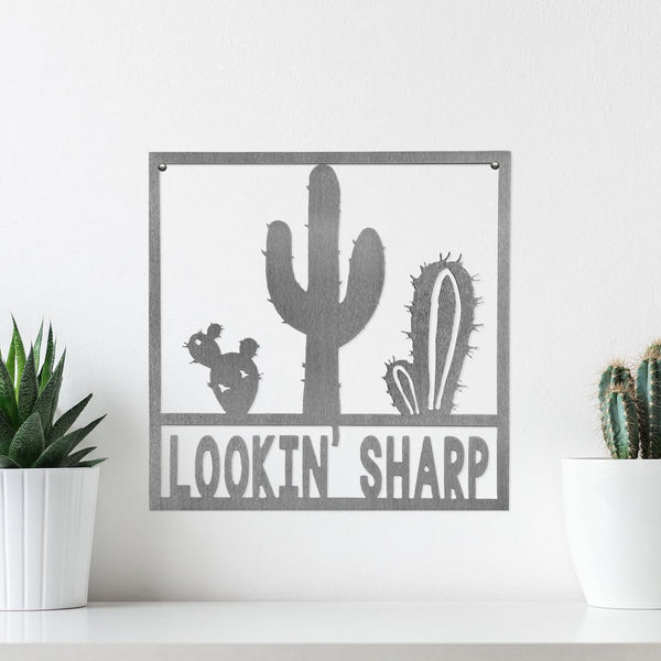 Square metal sign with cactus that says lookin sharp, hanging on wall.
