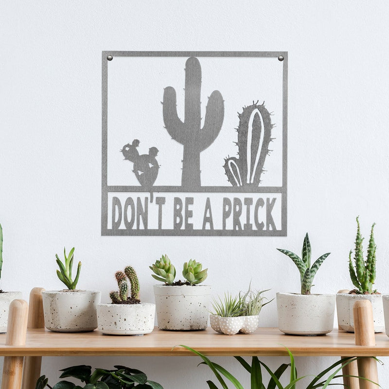 Square metal sign with cactus that says don't be a prick, hanging on wall above table.