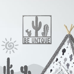 Square metal sign with cactus that says be unique, hanging on wall.