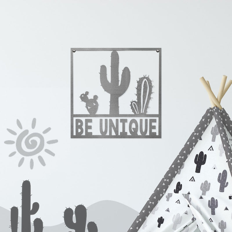 Square metal sign with cactus that says be unique, hanging on wall.