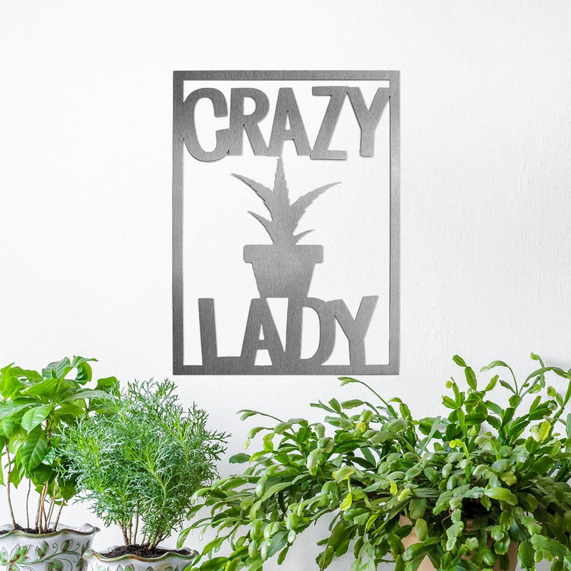 Rectangle metal sign that says crazy lady with a potted plant etched in the center, hanging above green plants.