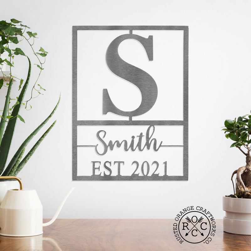 Personalized metal sign with monogram, name, and established date hanging on wall above plants.