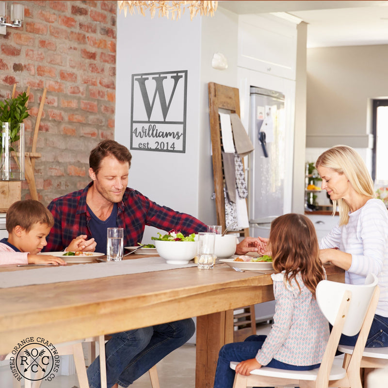 Personalized metal sign with monogram, name, and established date hanging on wall in kitchen, family eating around table.