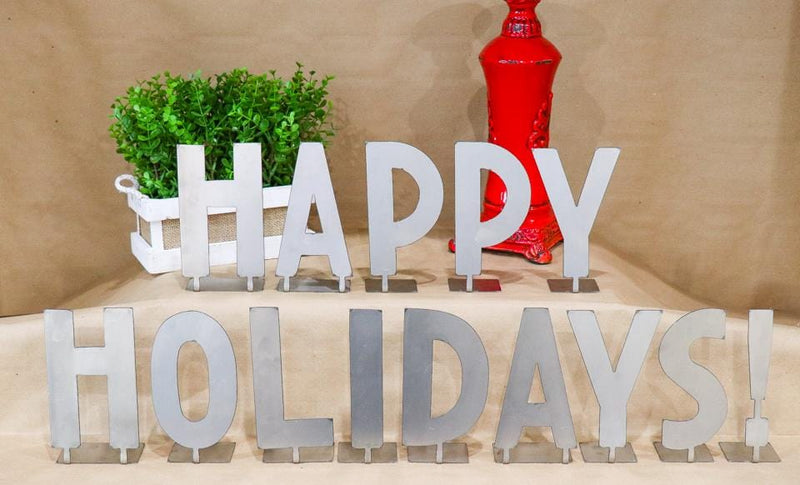 Metal letters with bent tabs on bottom spelling out happy holidays, standing on table next to plant and lamp.