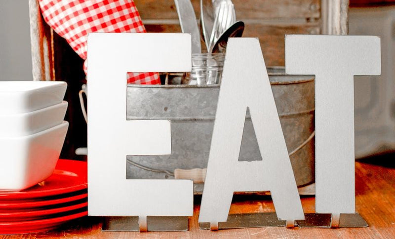 Metal letters with bent tabs on bottom spelling out the word eat, standing table next to dishes, silverware, and metal bucket.