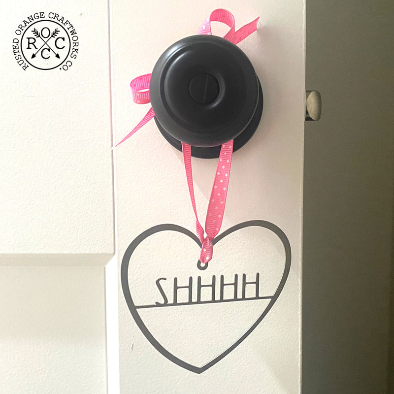 Heart shaped metal ornament customized with SHHHH hanging from doorknob with pink string.