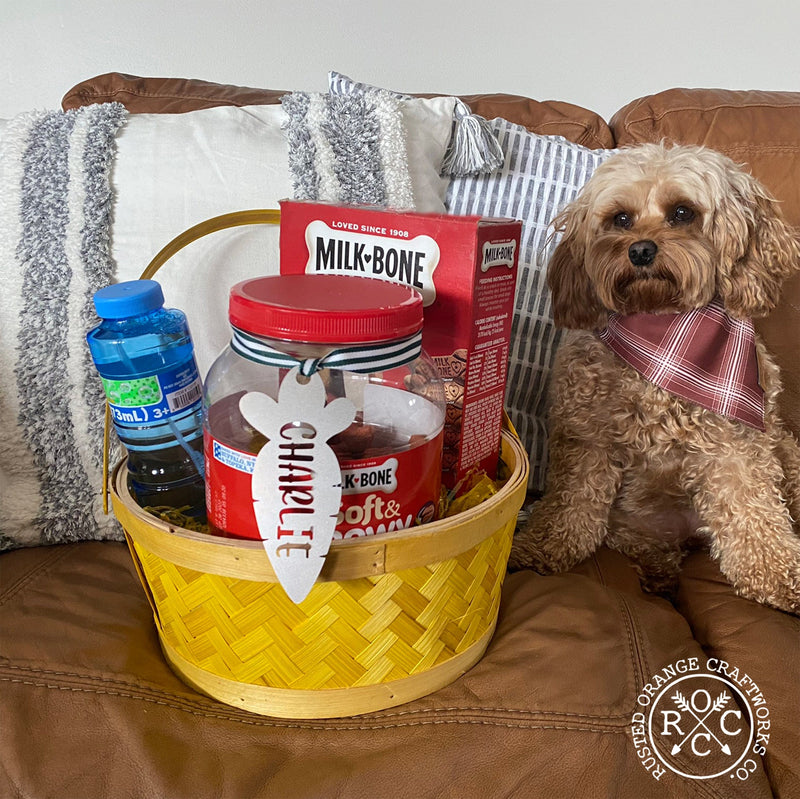 Dog sitting on couch next to Easter basket filled with dog treats, carrot shaped metal ornament with custom name of Charlie hanging from treats.