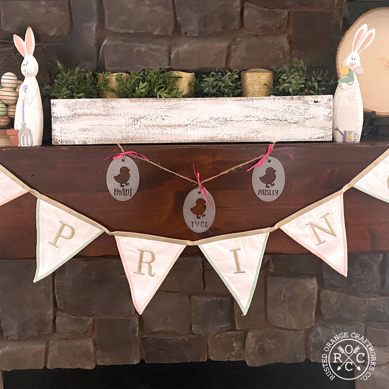 3 oval shaped metal ornaments with chick cutout and custom name below hanging on fireplace mantel.