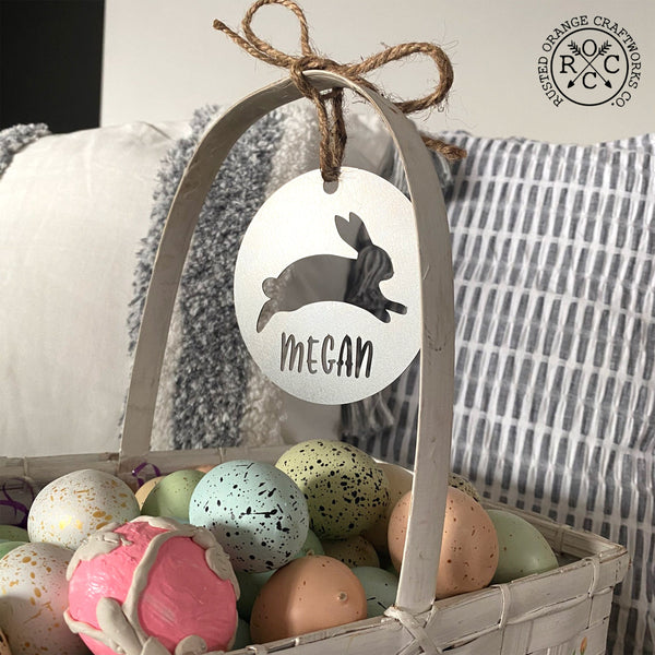 Easter basket filled with colored eggs, round metal ornament with bunny cutout in center and custom name of Megan below, tied to Easter basket handle.
