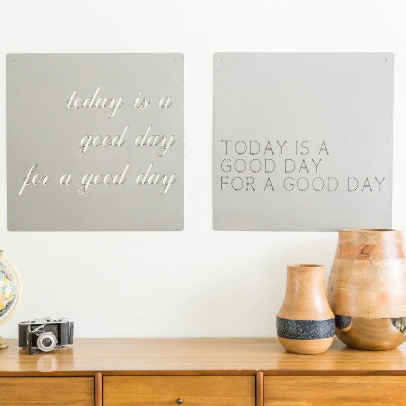 Two square metal signs saying today is a good day for a good day, hanging on wall above shelf.