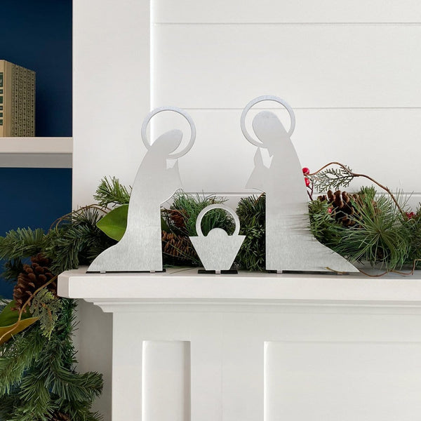 3 piece metal nativity scene silhouette standing on mantel in front of green garland.