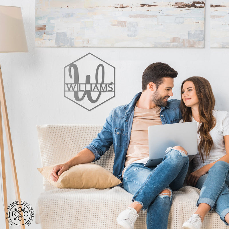 Hexagon shaped metal sign with last name and monogram in center, hanging on wall above couch, smiling couple sitting on couch.