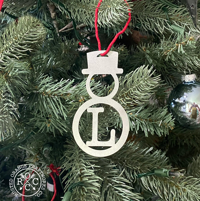 Metal snowman shaped ornament with monogram hanging from Christmas tree.