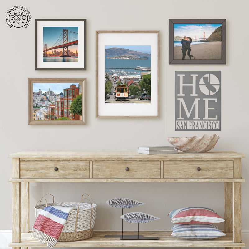 Metal sign saying home with home state silhouette in the oh and city name underneath, hanging on wall above shelf with photos.