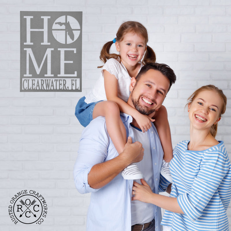 Metal sign saying home with home state silhouette in the oh and city name underneath, hanging on wall with smiling parents and daughter.