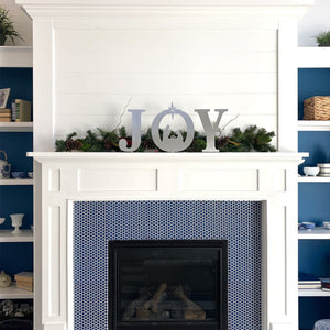 Metal letters J O Y with manger scene silhouette inside the o, standing on mantel in front of green garland.