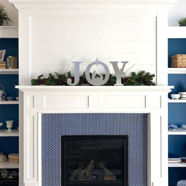 Metal letters J O Y with manger scene silhouette inside the o, standing on mantel in front of green garland.