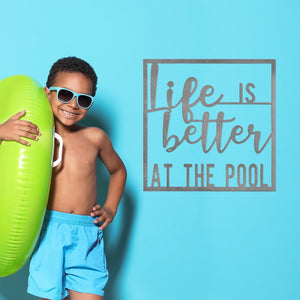 Square metal sign saying life is better at the pool, hanging on wall next to boy in swimsuit holding water toy.