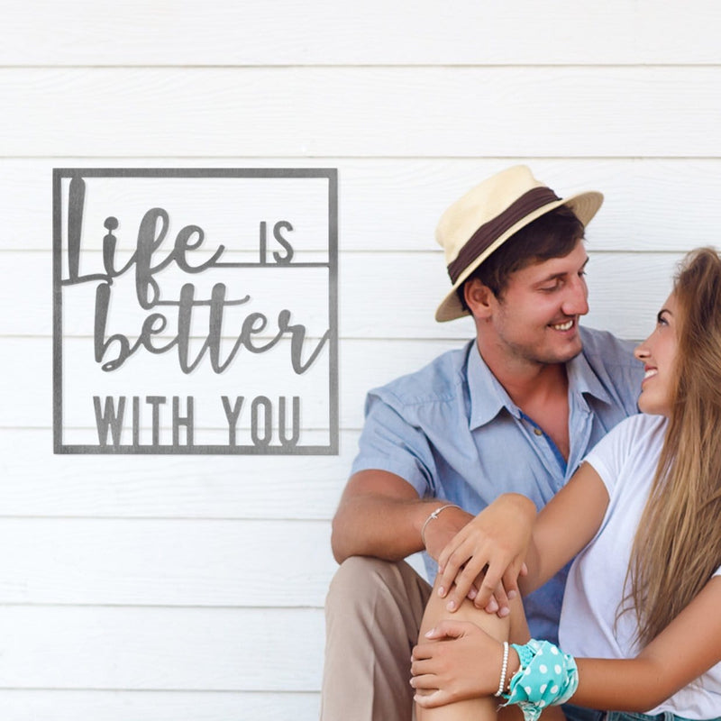 Square metal sign saying life is better with you, hanging on wall next to smiling couple.