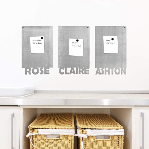 3 rectangle magnetic boards with names etched at the bottom with papers held by magnets on them, hanging on wall above shelf.
