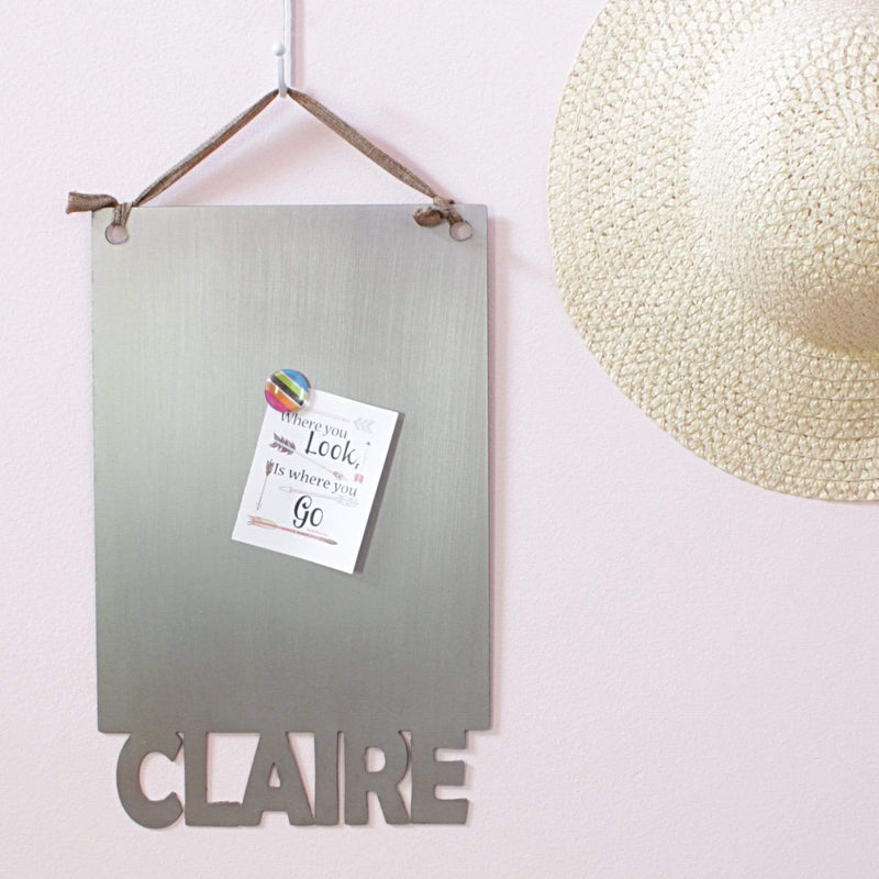 Rectangle magnetic board with name etched at the bottom with paper held by magnet, hanging on wall next to straw hat.