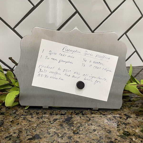 Small metal board with recipe stuck on with magnet, sitting on countertop next to flowers.