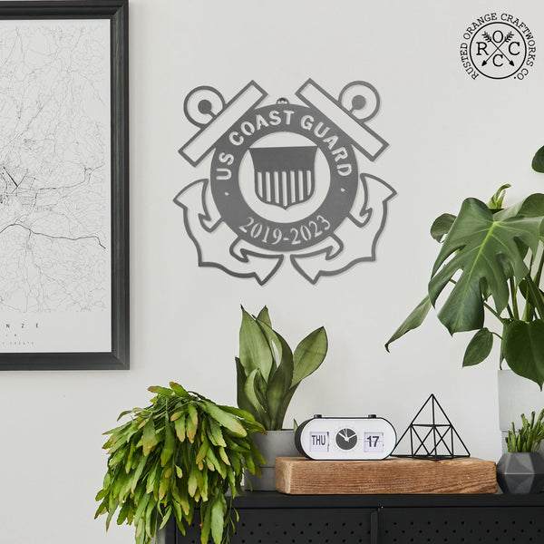 Round metal sign saying US Coast Guard along top and years of service on bottom with 2 anchors behind and coast guard symbol in the center, hanging on the wall above shelf with plants.